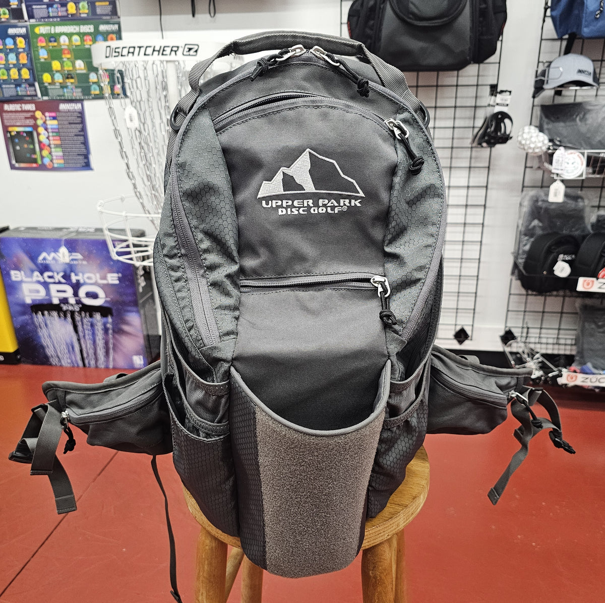 A dark gray and black disc golf backpack with a white 'Upper Park Disc Golf' logo on the front, displayed on a wooden stool inside a retail store.